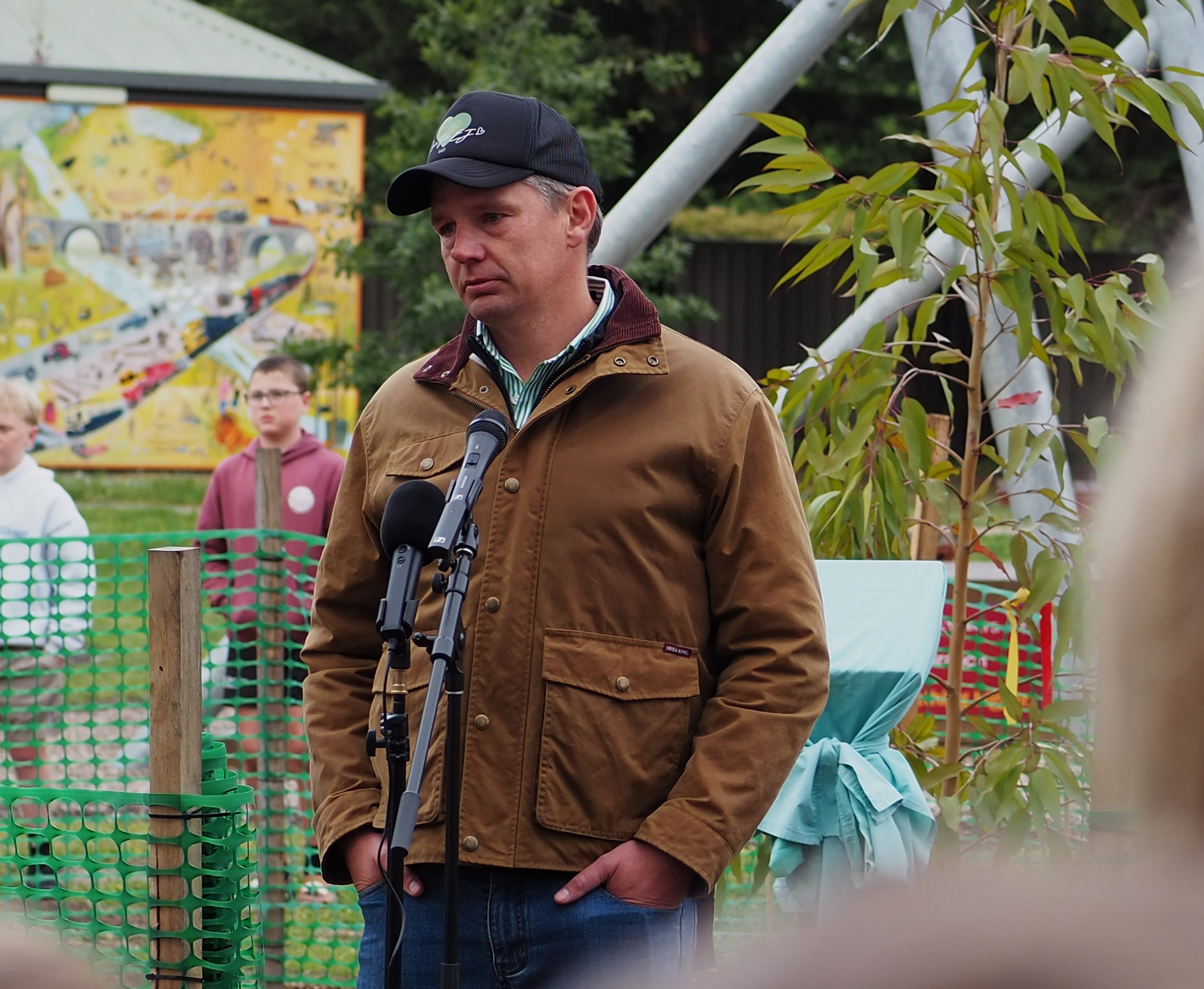 Um homem de jaqueta marrom, rosto carrancudo e mãos nos bolsos, fala para a multidão.