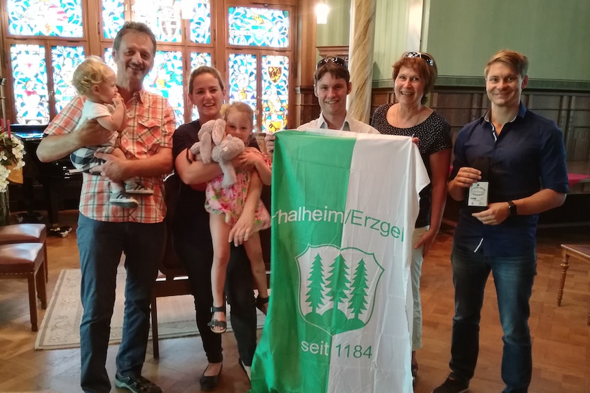 Five adults and two children posing with a town flag displaying German writing.