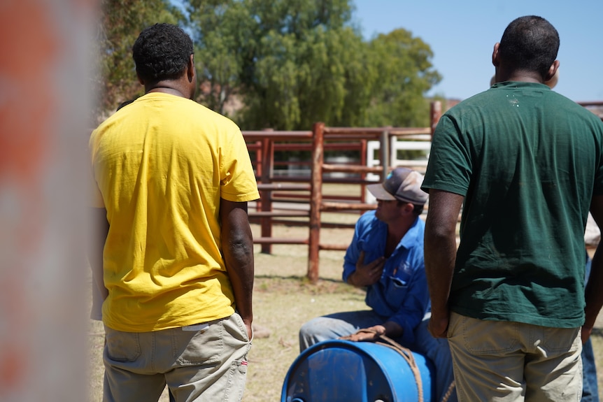 Two prisoners stand on either side of a man wearing a blue shirt sitting astride a barrel. Their backs are to the camera.