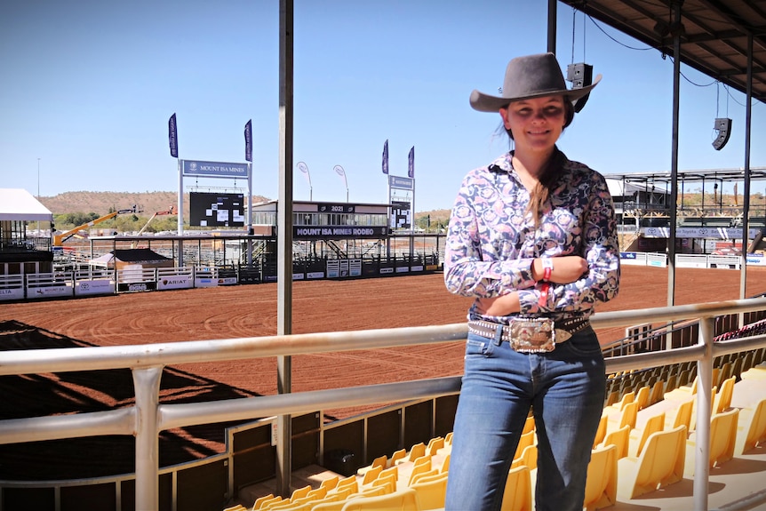 A girl wearing rodeo outfit stands in the Mount Isa Rodeo grounds with the arena behind her.