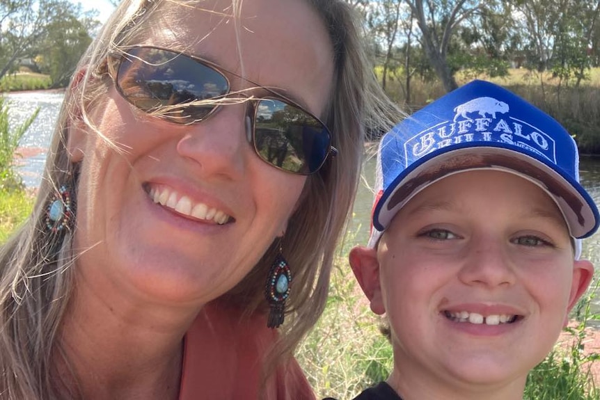 Woman with sunnies and blonde hair smiles at camera next to little boy wearing blue cap