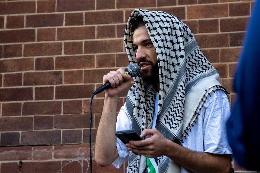 Man wearing white t-shirt, with a keffiyeh over his head, holding microphone.