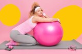 A white blonde woman rests on a gray exercise mat with a pink exercise ball in front of her that she leans on while pouting.