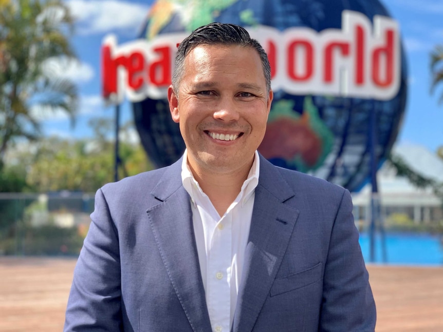 A man wearing a grey suit smiles at the camera with a Dreamworld sign behind him