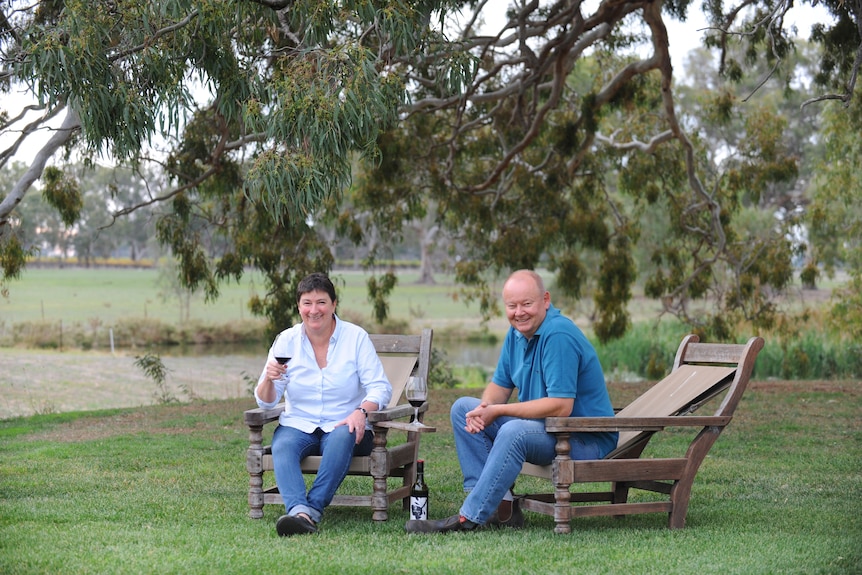 A woman with brown hair and a white shirt and a man in a blue short and blue jeans sit on chairs in a winery