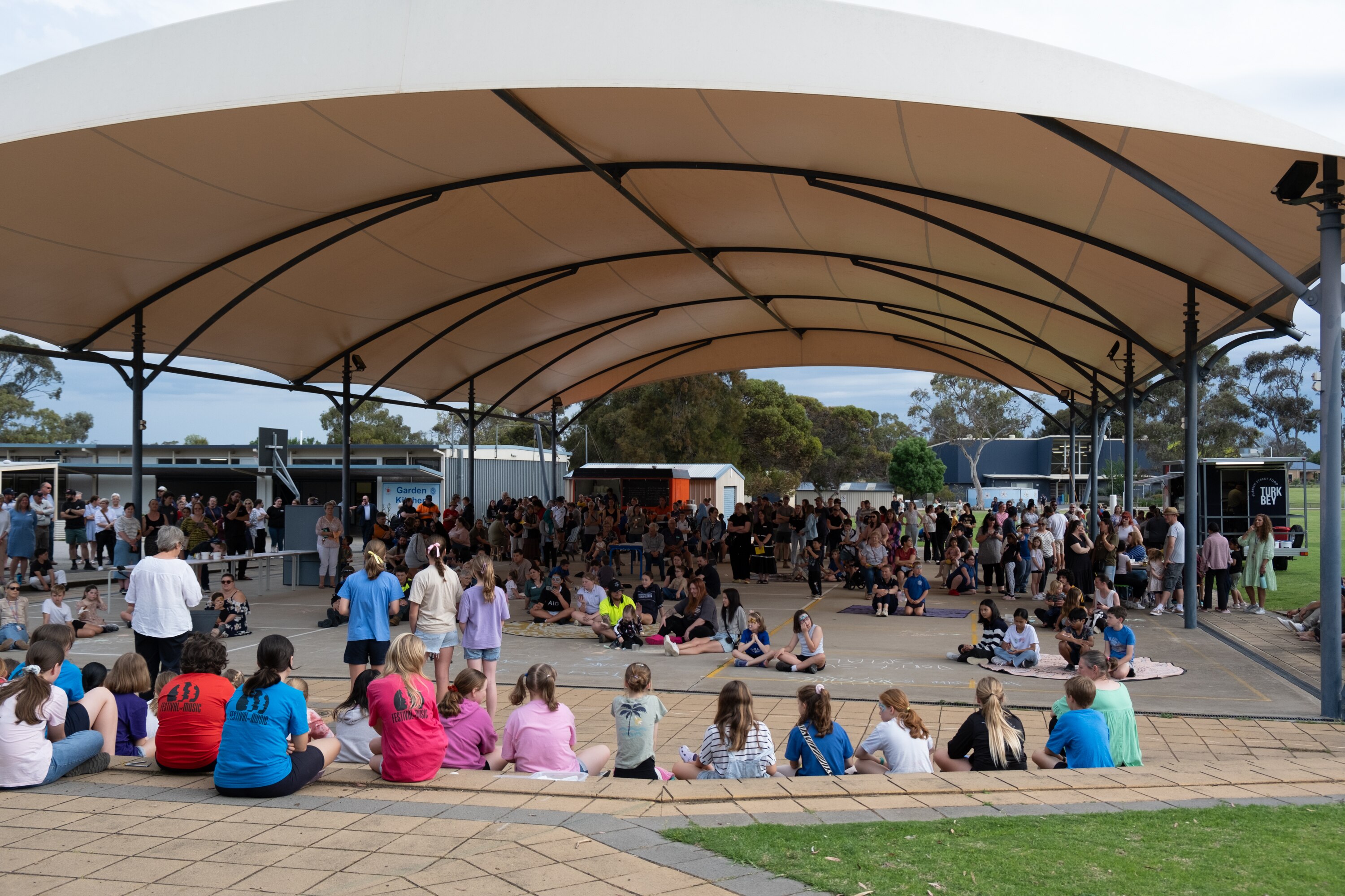 A woman speaks to a crowd of children and adults under a shade shelter