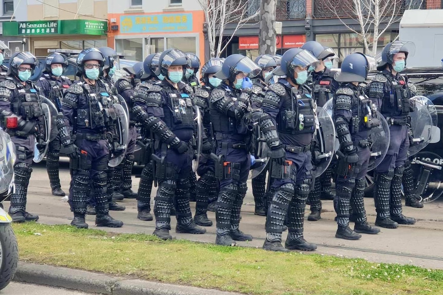 A large police presence in riot gear, all wearing surgical masks, stands on a Melbourne street.