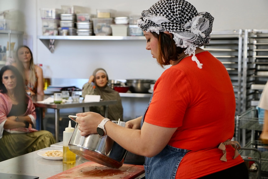 A woman works in a kitchen while others watch intently.
