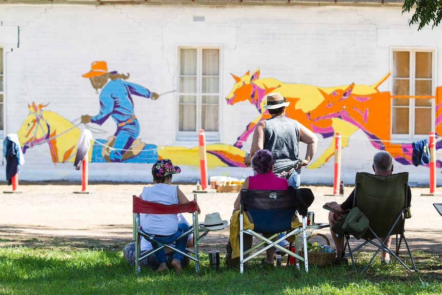 Three people sitting in chairs on grass watching a mural being painted on a building.