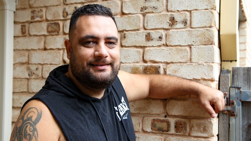 A New Zealand man with a tattoo on his arm leans against a brick wall.