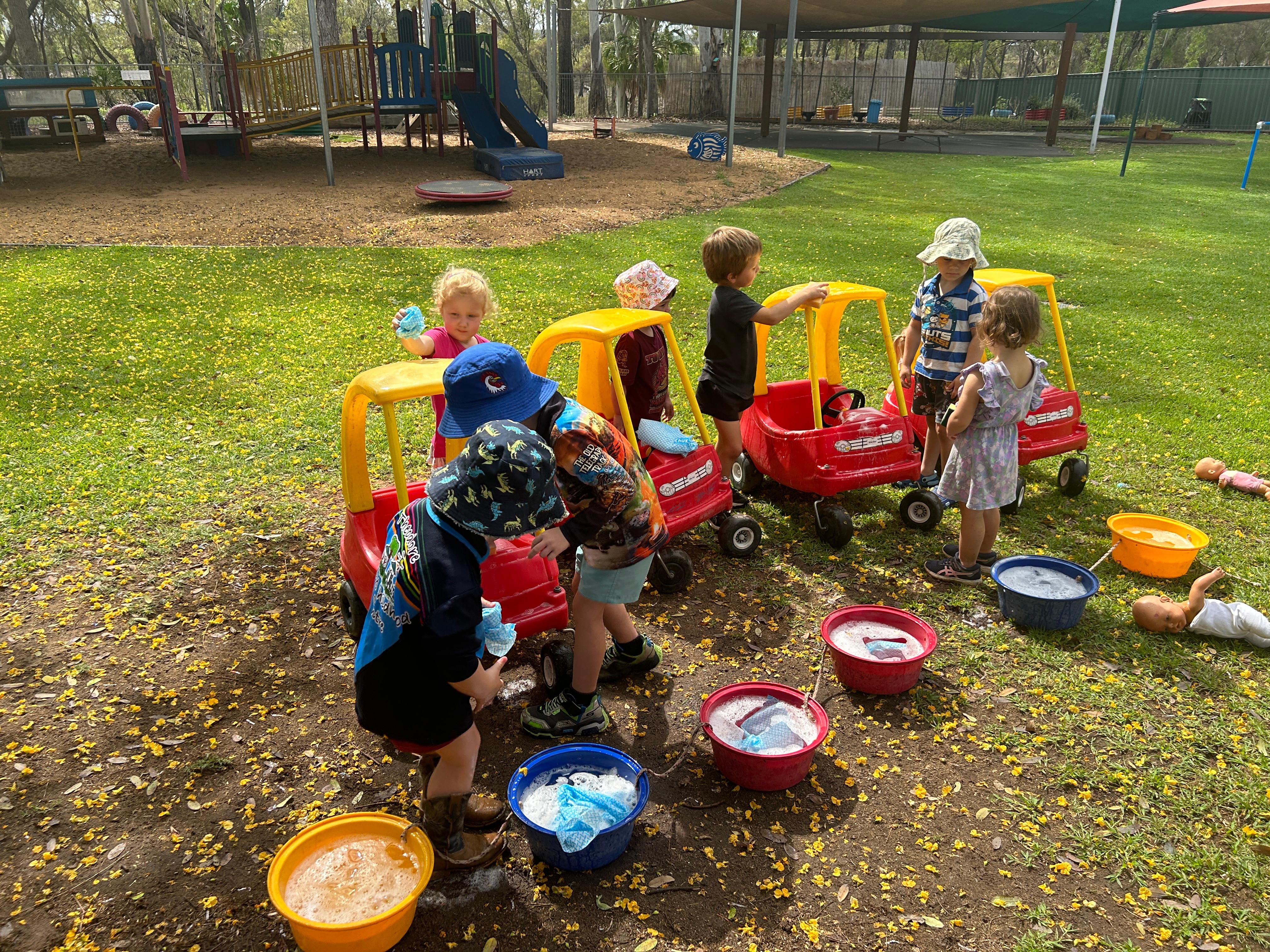 A group of kids play child sized large plastic cars and water buckets on grass in front of a large playground  