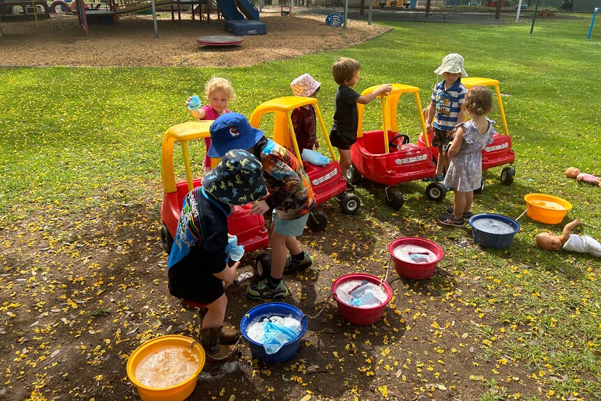 A group of kids play child sized large plastic cars and water buckets on grass in front of a large playground  