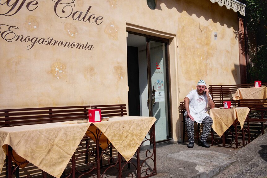 A man in a chef's uniform sits talking on the phone outside a restaurant