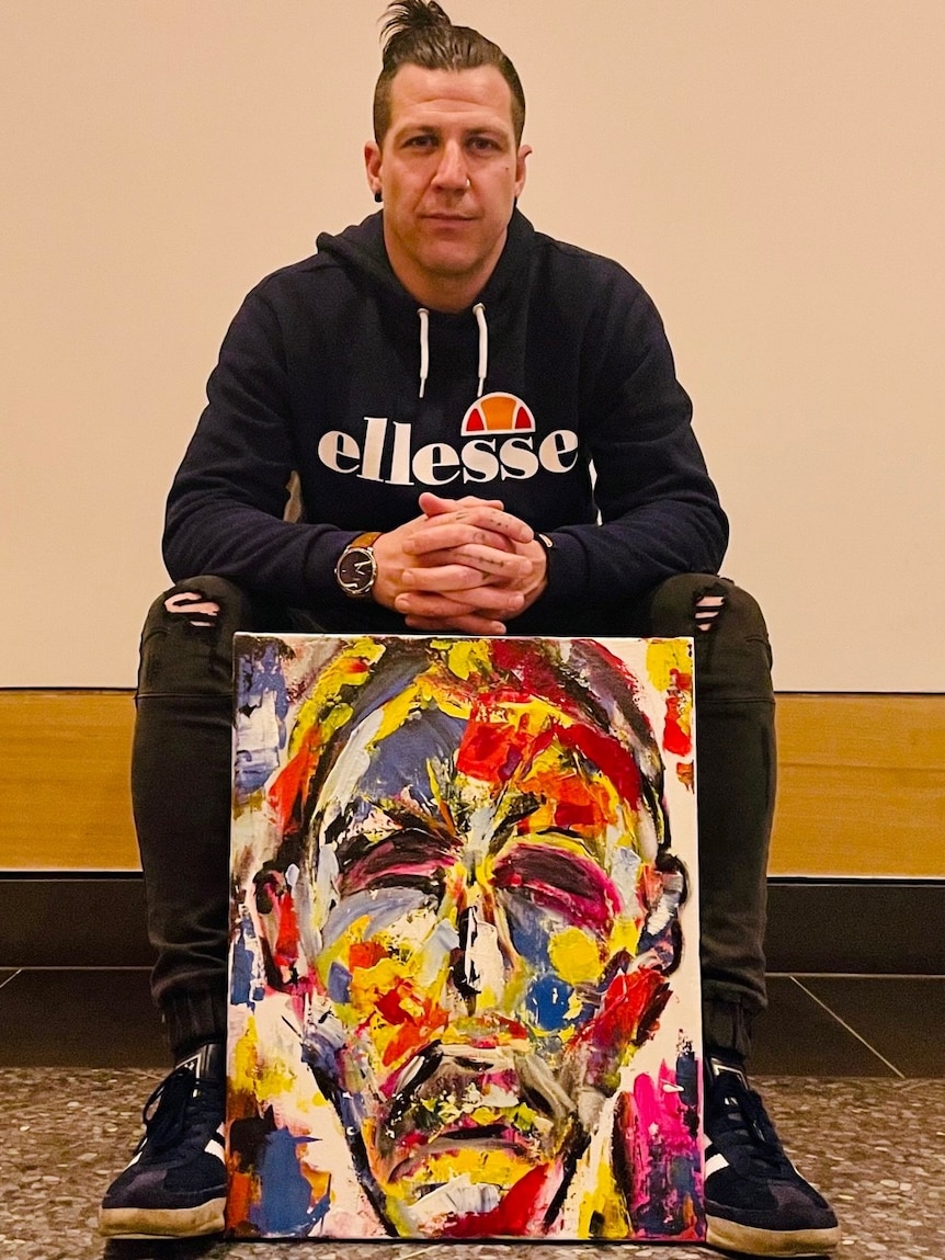 A man in a blue hoody and hair pulled back in a bun, sits on a seat with an acrylic self-portrait resting against his knees. 