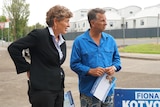 Woman in black suit looking left, standing nect to man looking left wearing blue shirt and holding piece of paper