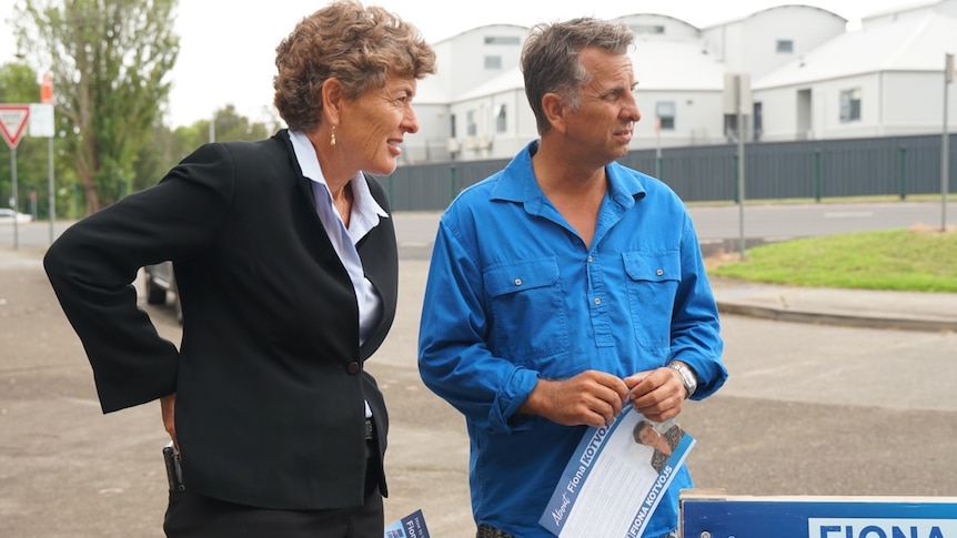 Woman in black suit looking left, standing nect to man looking left wearing blue shirt and holding piece of paper
