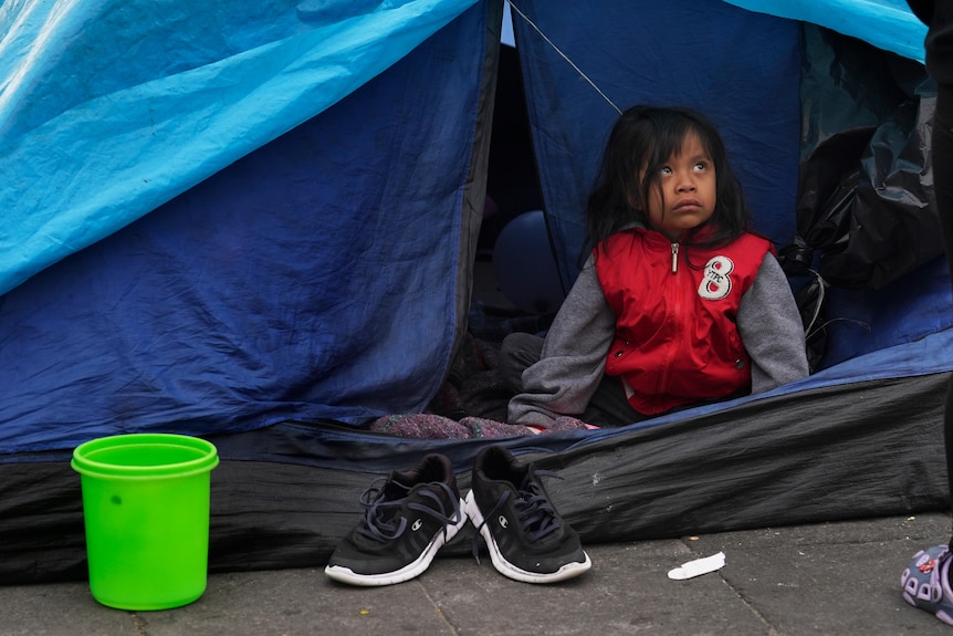 A young girl in a red jacket looks forlornly out of a blue tent on concrete.