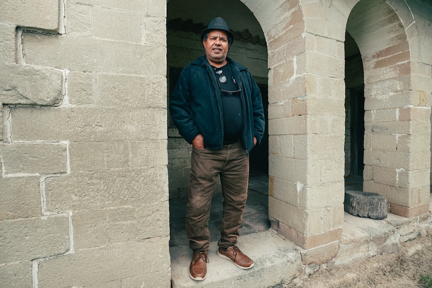 An Indigenous man wearing a dark hat and jacket standing in a stone archway at a historic homestead.