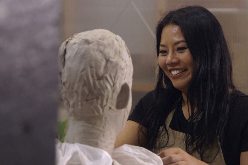 The sculptor, with long black hair, sits with a small clays study in the shape of a human head sitting in front of her.