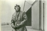 A historic photo of an aviator standing outside a aircraft hanger with flight googles on his head.