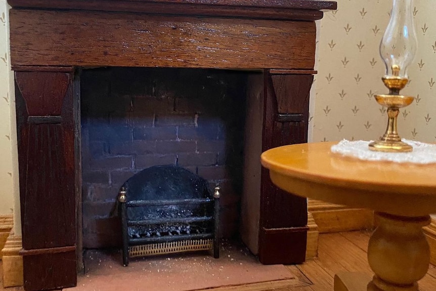 Photograph of a miniature fireplace and grate, with a round table and kerosene lamp on top.