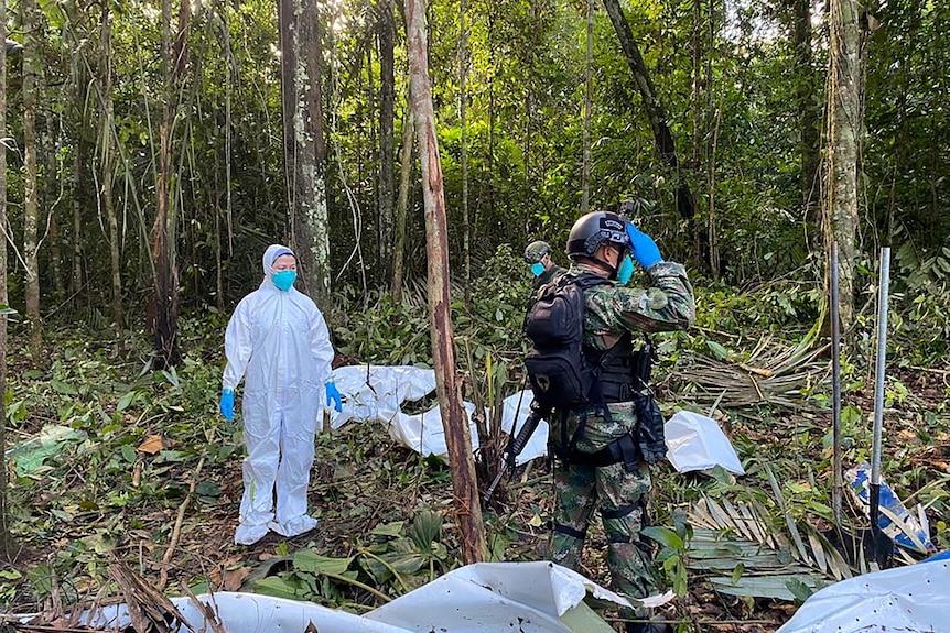 A person in a hazmat suit and another in army gear standing in a forest surrounded by plane debris. 