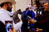 A man with a beard and cowboy hat holds up a grey, hairless cat for journalists who take pictures with phones and cameras.