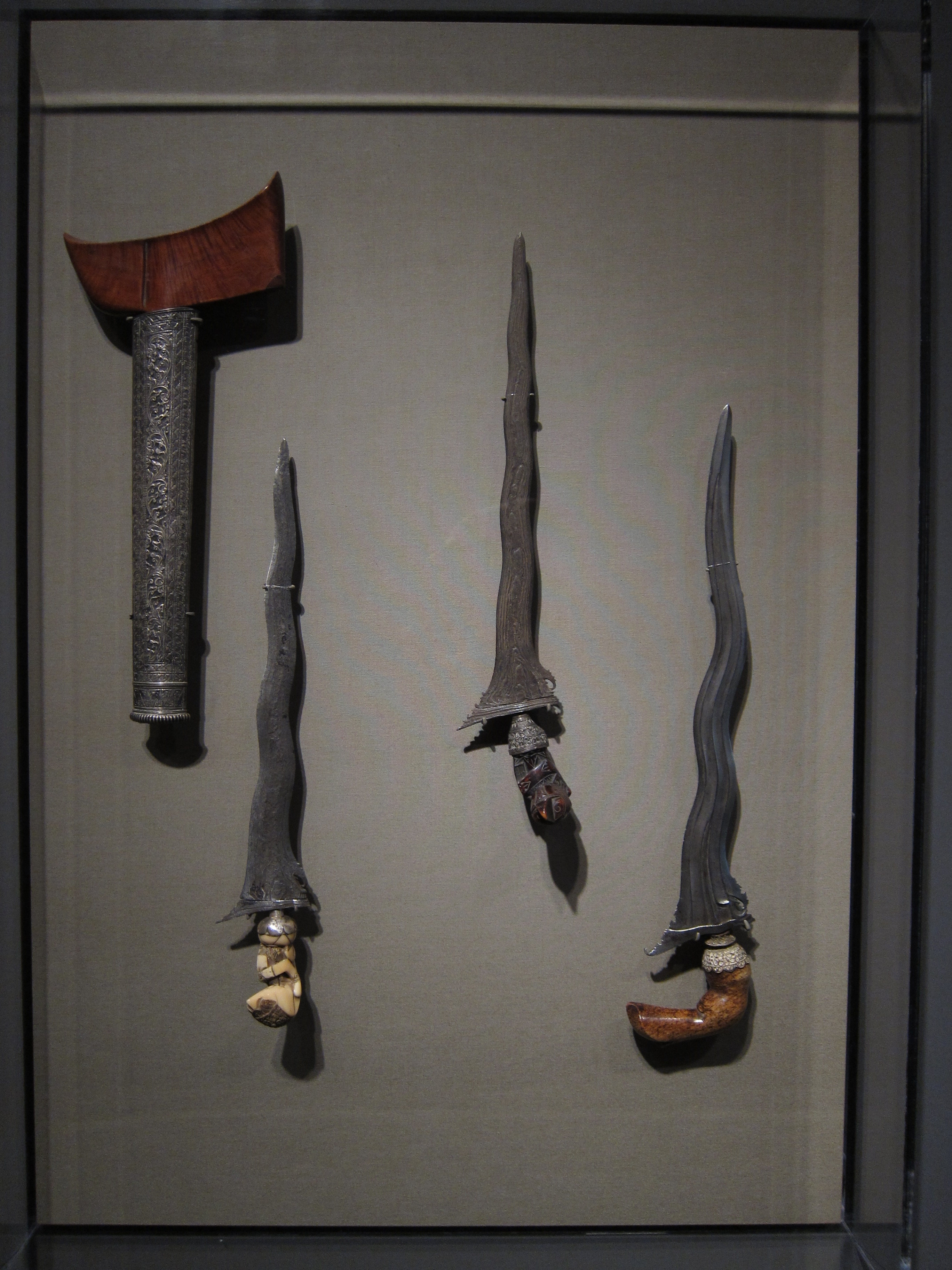 Three wavy-bladed daggers and a metal and wooden sheath mounted in a frame.