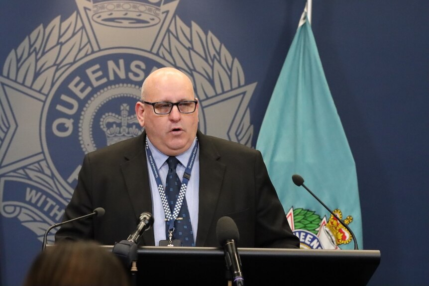 A man in a black suit standing in front of a Queensland Police Service logo talks to reporters
