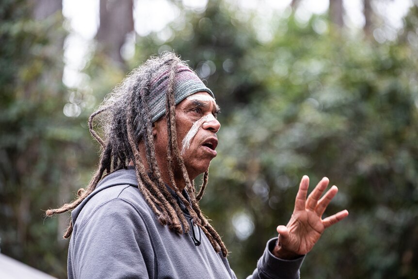 An Indigenous man with paint on his face and long dreadlocks gestures as he speaks in a forest clearing.