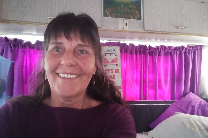 A woman smiling in a caravan, with a magenta curtain across the window. 