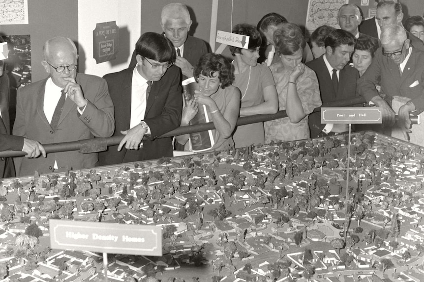 A black and white image of a people crowding around table with a model of a town