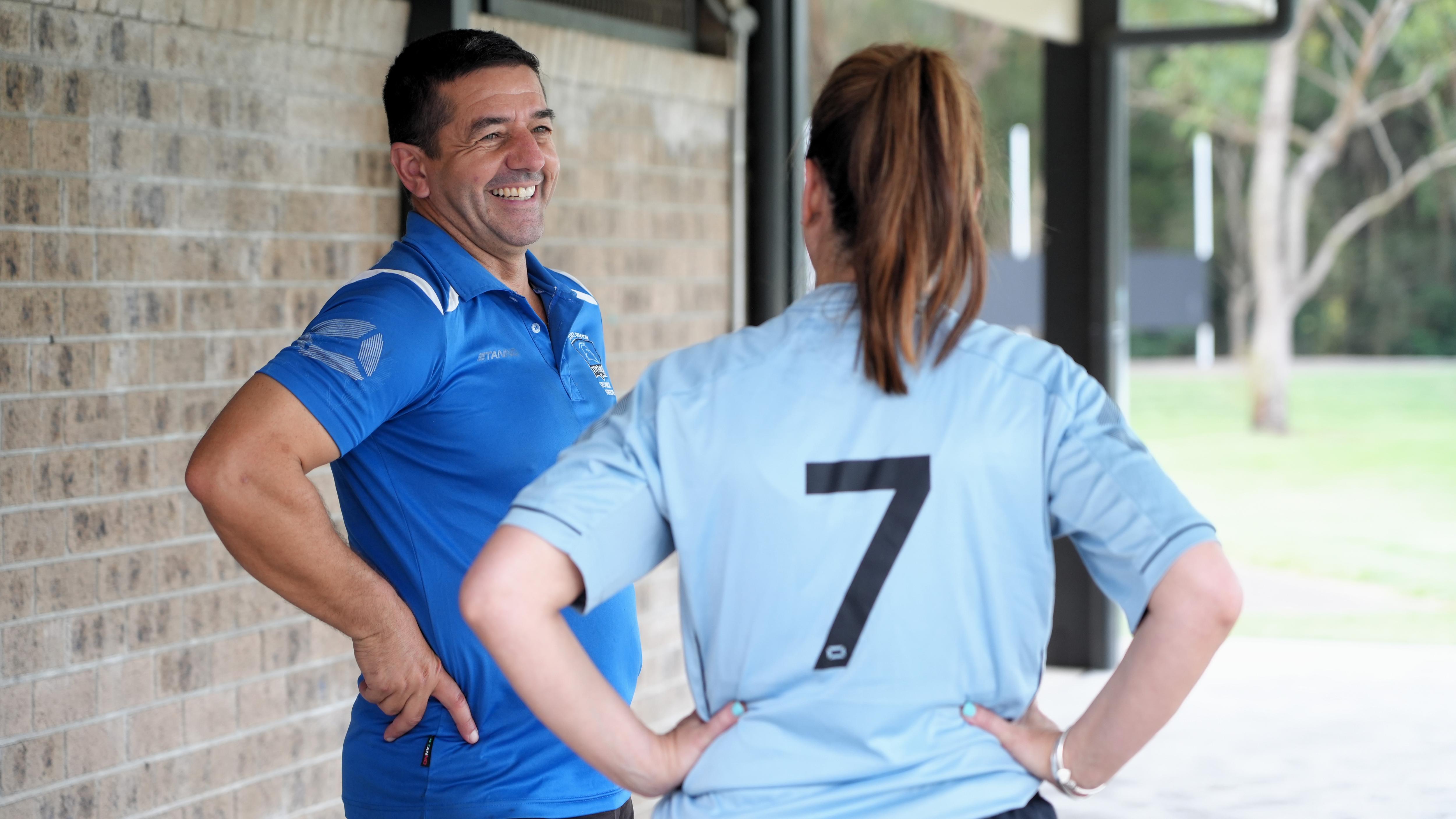 Um homem vestindo uma camisa pólo azul tem as mãos nos quadris e sorri enquanto fala com uma jogadora de futebol