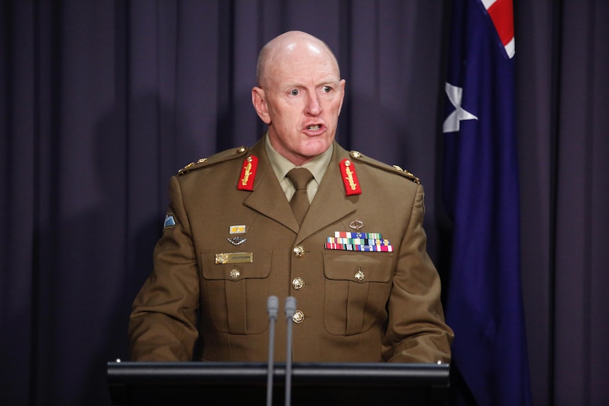 Frewen, un homme chauve, porte un uniforme militaire de l'armée debout derrière des microphones et devant un drapeau australien.