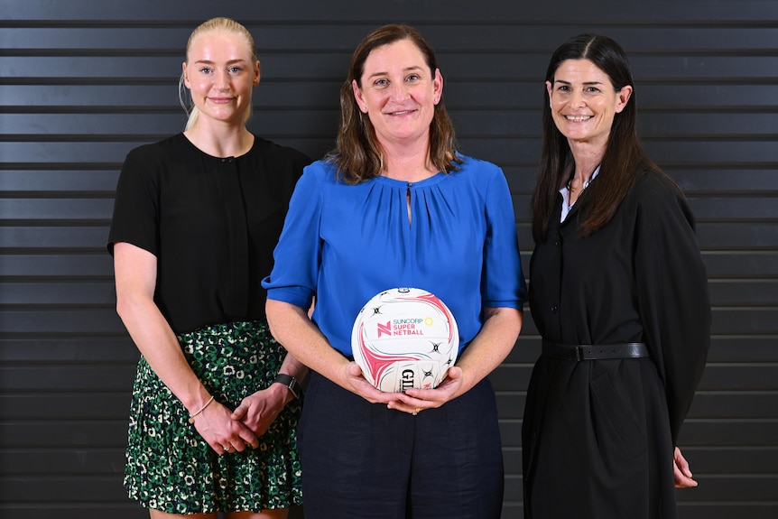 Three women stand together, as the acting netball australia ceo holds a ball in the middle