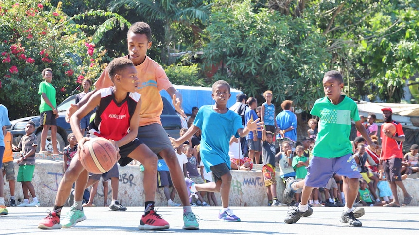 A young player holds the ball looking for a teammate on an outdoor basketball court with a crowd and trees in the background
