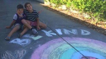 Two boys with their arms around each other sit in front of a chalk drawing saying be kind.