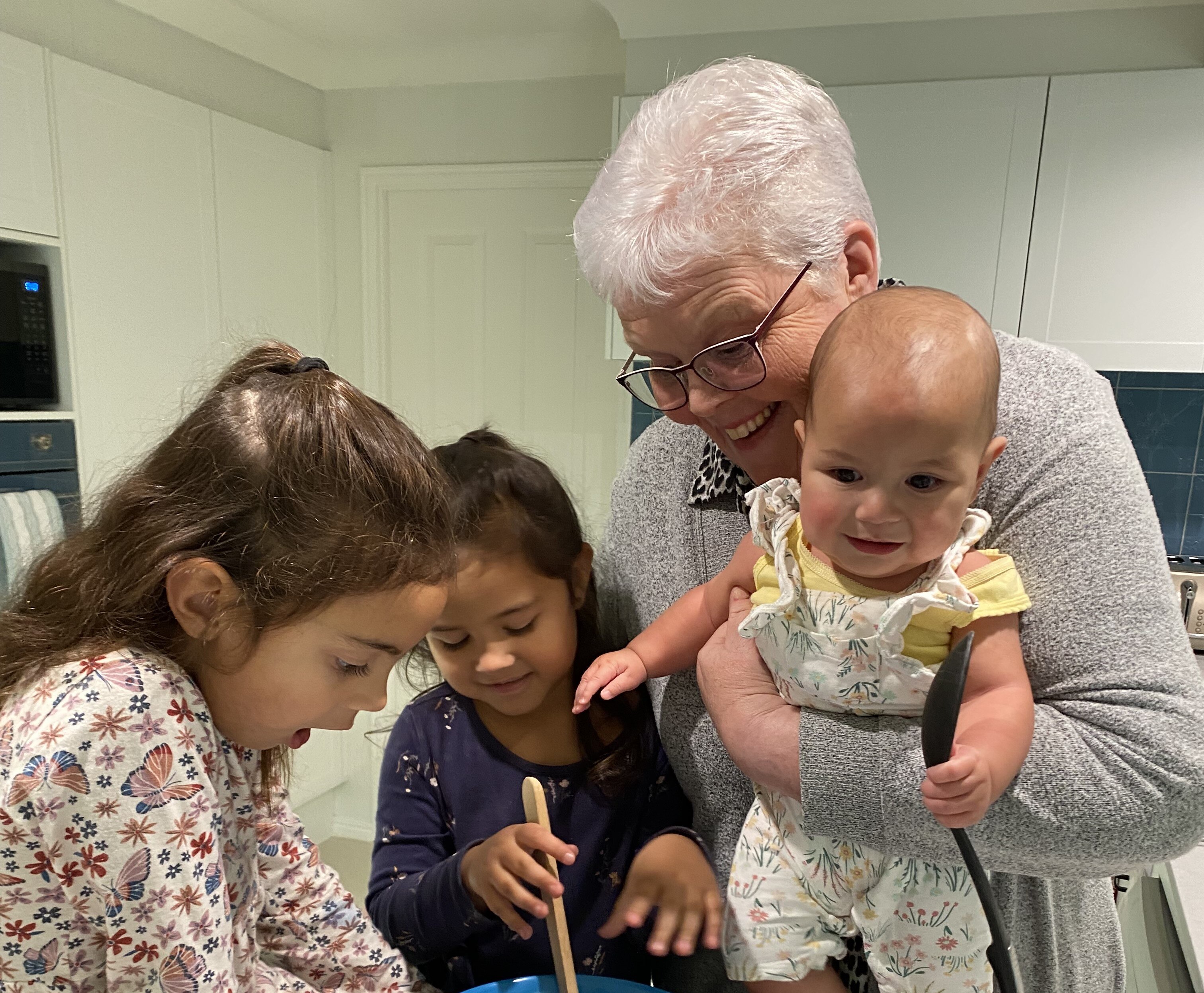 An older lady on the right of the frame holds a baby and watches over two young girls cooking