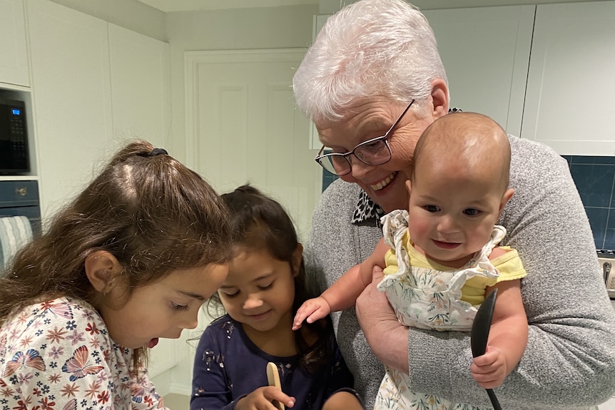 An older lady on the right of the frame holds a baby and watches over two young girls cooking