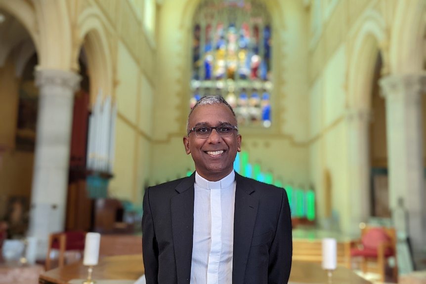 A smiling South Asian man wearing priest's collar, dark suit jacket, stands in a building with arches and stained glass .