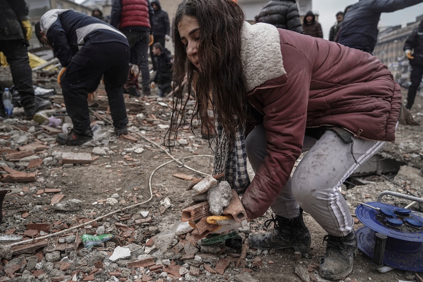 A woman with long, dark hair picks up handfuls of rubble at the site of a collapsed building.