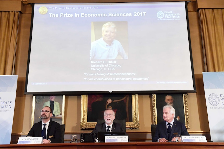A panel of men announcing the Nobel Economics Prize and a projection of the winner behind them.