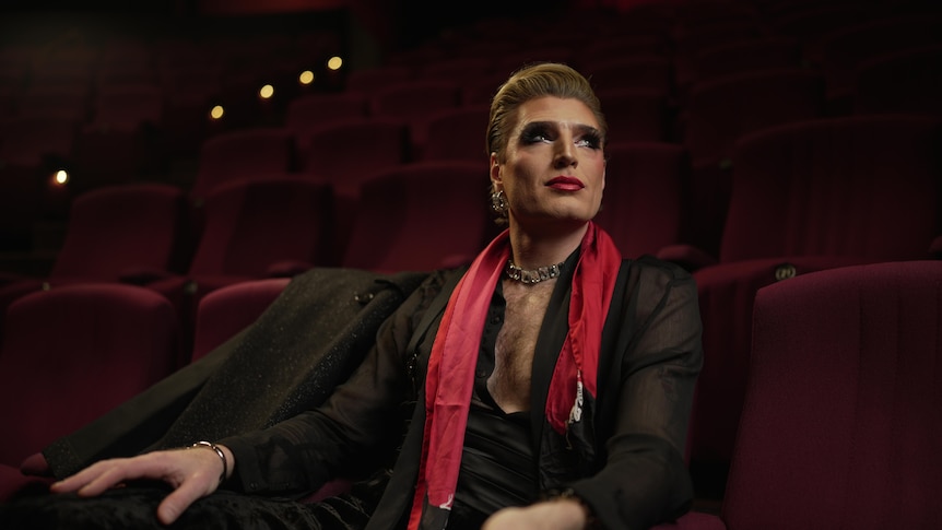 A man in his 30s in drag makeup, in a black sheer shirt and red scarf, sitting in dark red audience seats