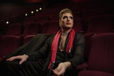 A man in his 30s in drag makeup, in a black sheer shirt and red scarf, sitting in dark red audience seats