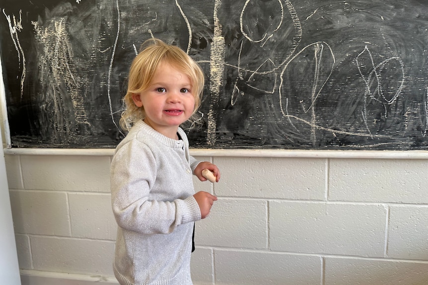 A cute two-year-old girl smiles at the camera in front of a black board.