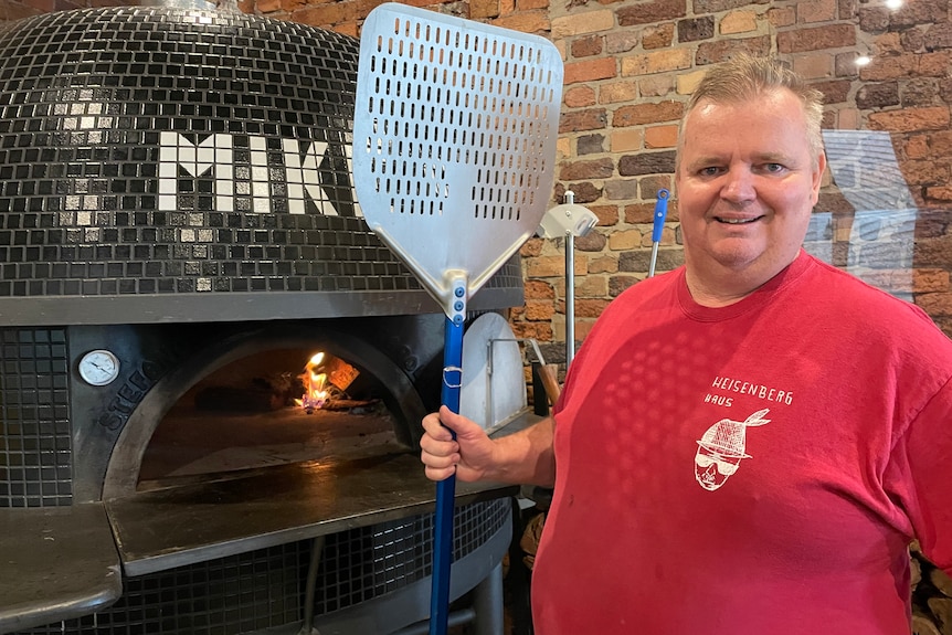 Pizza shop owner stands next to a pizza oven.