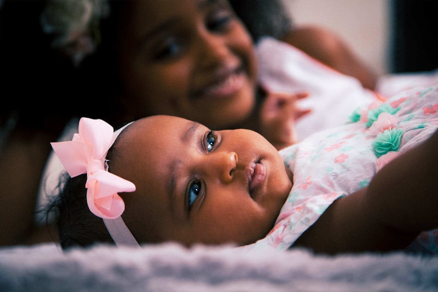 Very cute baby girl wearing a pink bow with her mum blurred in the background, for a story about unique baby name popularity.