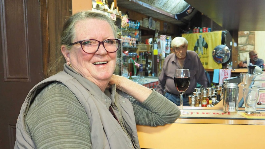 Woman sits at a bar with a glass of red wine as a man stands behind the bar in the background.