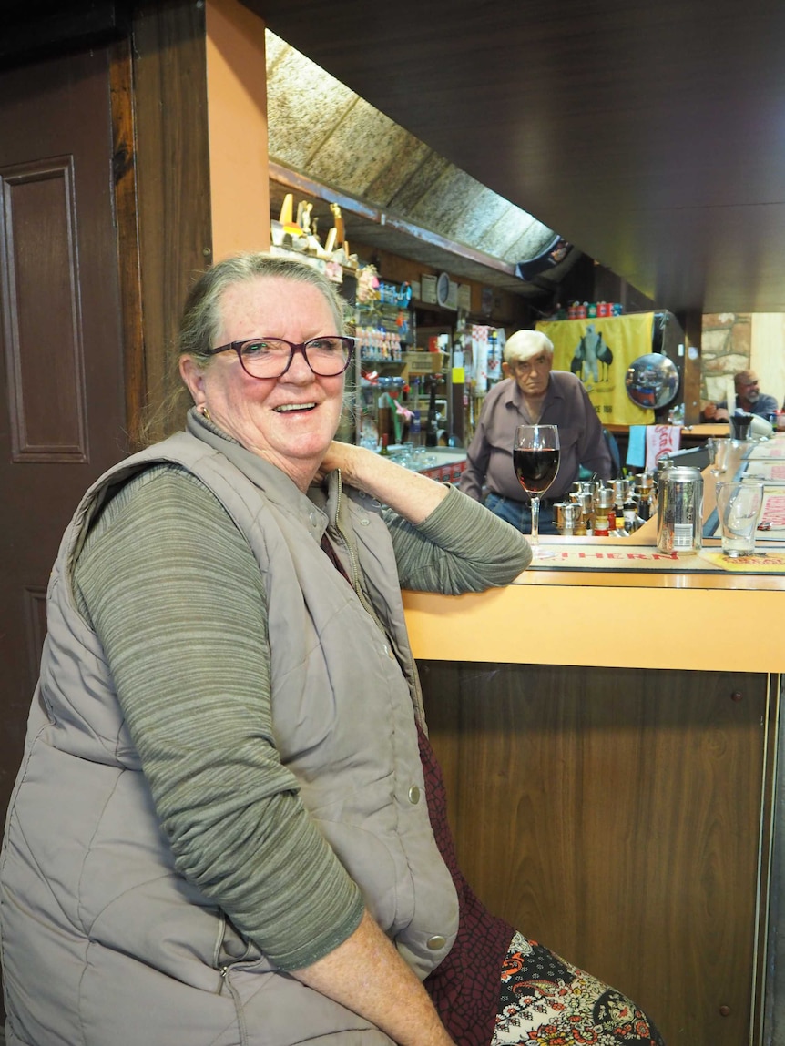 Woman sits at a bar with a glass of red wine as a man stands behind the bar in the background.