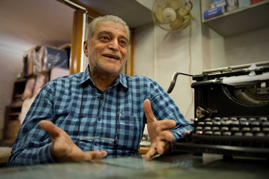 An older man in a blue checked, button up shirt, sits at a table next to a typewrite, gesturing, mid-conversation